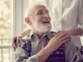 Ein älterer Mann im Rollstuhl lächelt, während eine Pflegekraft ihm die Hand auf die Schulter legt. Er fragt sich, wie lange es dauert, bis der medizinische Dienst kommt.