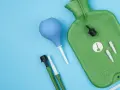 Grüne Wärmflasche mit Zubehör für einen hebe schwenkeinlauf. Blaue Birne und grüne Schläuche mit Aufsätzen liegen daneben.