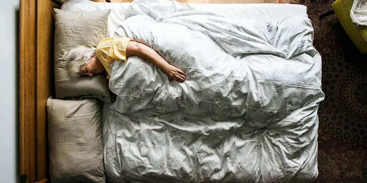 Eine ältere Frau schläft tief und fest in einem gemütlichen Bett. Wenn alte Menschen viel schlafen, ist das oft ein Zeichen von Ruhe und Erholung.