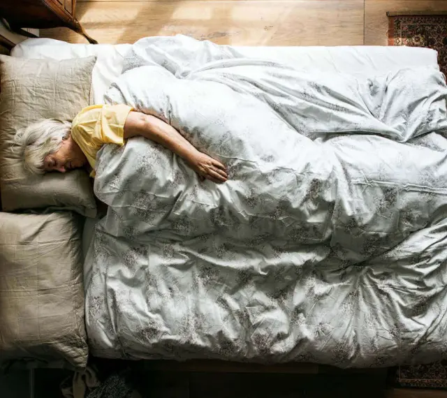 Eine ältere Frau schläft tief und fest in einem gemütlichen Bett. Wenn alte Menschen viel schlafen, ist das oft ein Zeichen von Ruhe und Erholung.