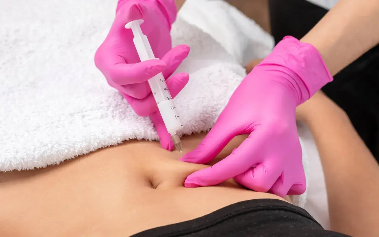 Hände in pinken Handschuhen führen eine subkutane Injektion im richtigen Winkel am Bauch durch.
