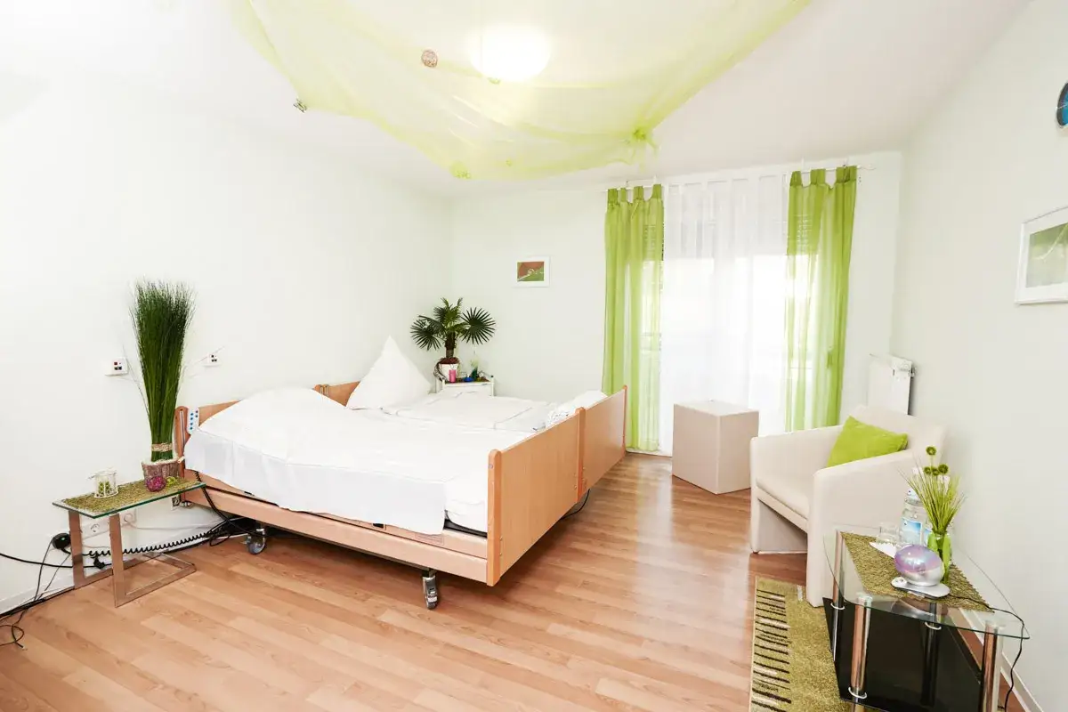 Ein helles Zimmer mit einem Pflegebett, das für wachkoma patienten geeignet ist. Ein Sessel und ein Nachttisch vervollständigen die Einrichtung.