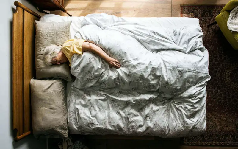 Eine ältere Frau schläft tief und fest in einem gemütlichen Bett. Wenn alte Menschen viel schlafen, ist das oft ein Zeichen von Ruhe und Erholung.